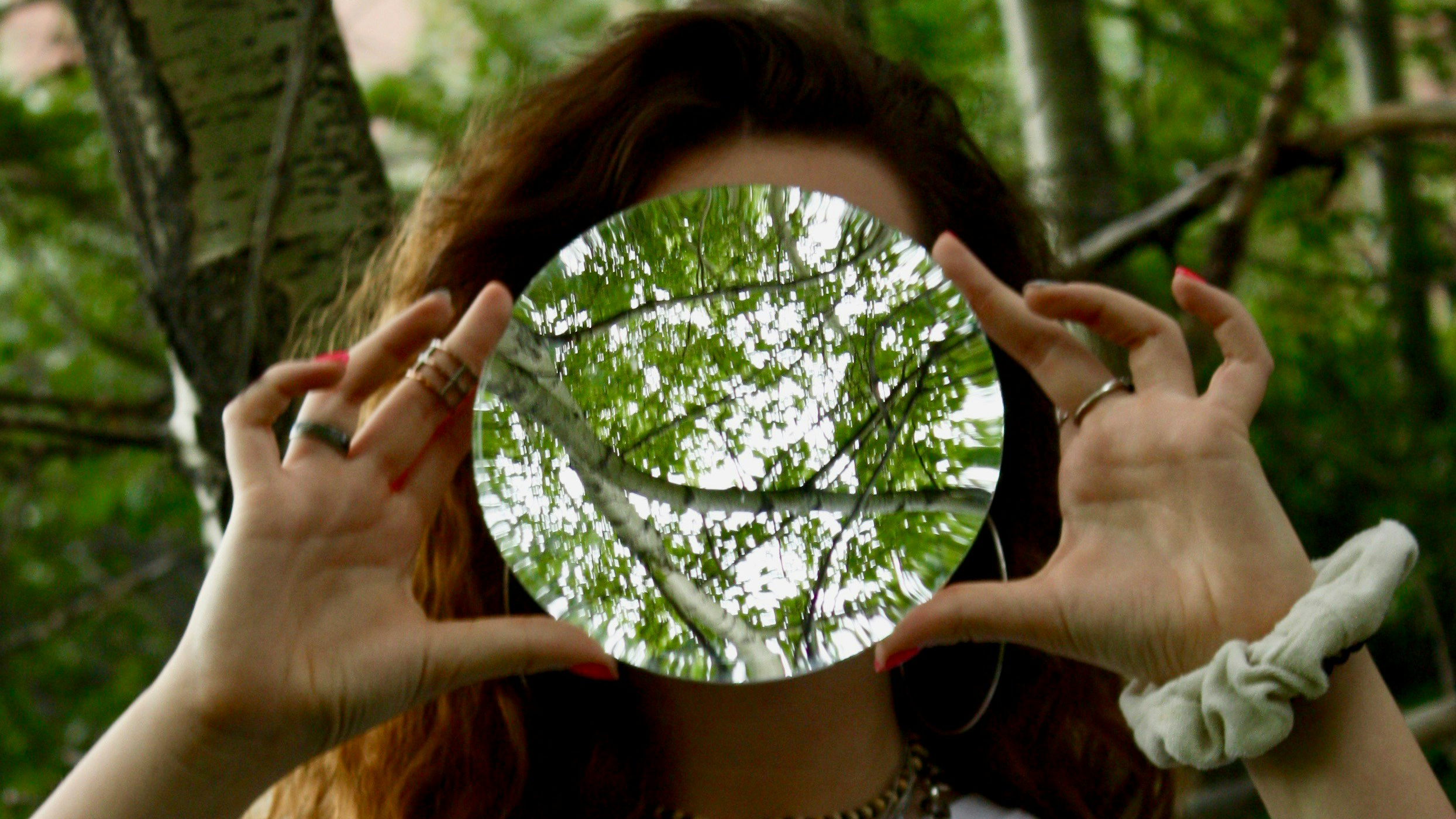 Woman standing under a tree with a circular mirror in front of her face reflecting back more trees.